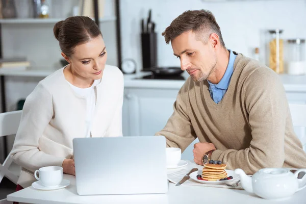 Adult couple sitting at table and using laptop during breakfast in morning — Stock Photo