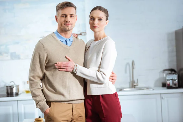 Erwachsenes Paar in lässiger Kleidung blickt in die Kamera und umarmt sich in der Küche — Stockfoto