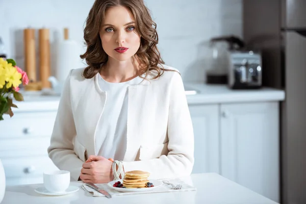 Belle femme assise à table avec crêpes et café pendant le petit déjeuner dans la cuisine — Photo de stock