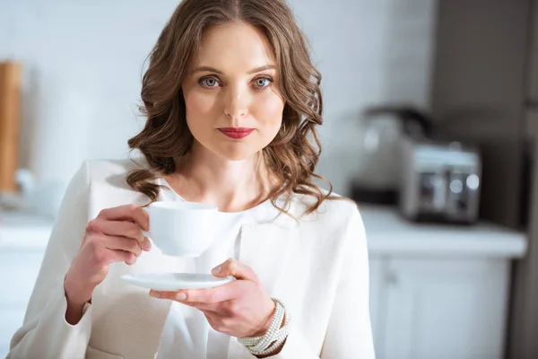 Bela mulher segurando xícara de café e olhando para a câmera durante o café da manhã na cozinha — Fotografia de Stock