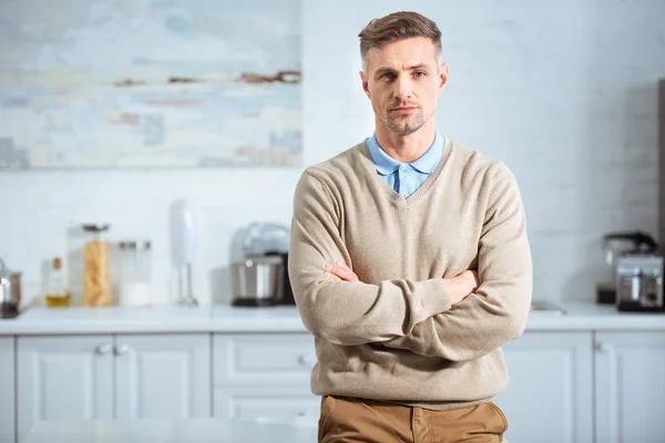 Hombre guapo en ropa casual con los brazos cruzados mirando a la cámara en la cocina - foto de stock