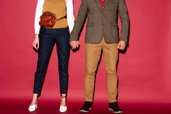 Vue recadrée du couple à la mode en tenue formelle tenant la main et posant sur fond rouge — Photo de stock