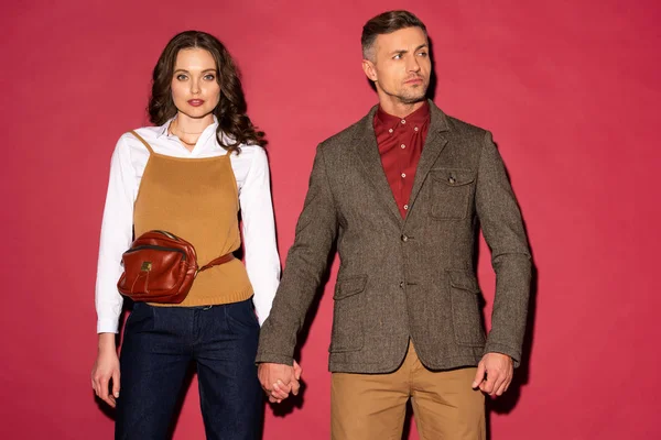 Bella coppia alla moda in abbigliamento formale che si tiene per mano e posa su sfondo rosso — Foto stock