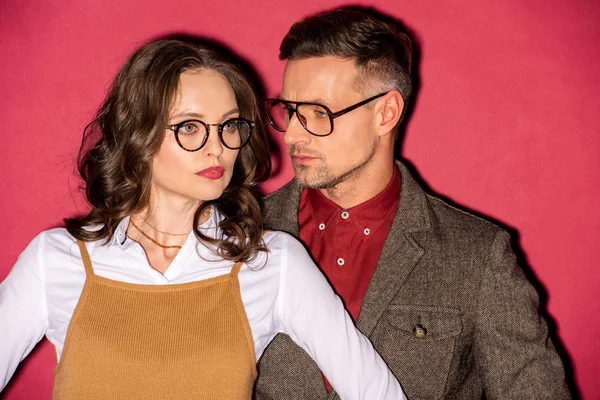 Belo casal na moda em desgaste formal e óculos posando no fundo vermelho — Fotografia de Stock