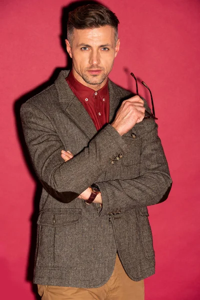 Handsome man in formal wear looking at camera and holding glasses on red background — Stock Photo