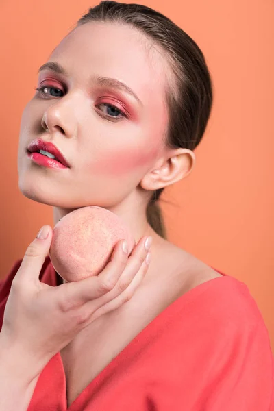 Beautiful fashionable girl holding peach, looking at camera and posing isolated on living coral — Stock Photo