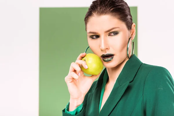 Hermosa mujer elegante sosteniendo manzana, mirando hacia otro lado y posando con verde marino en el fondo - foto de stock