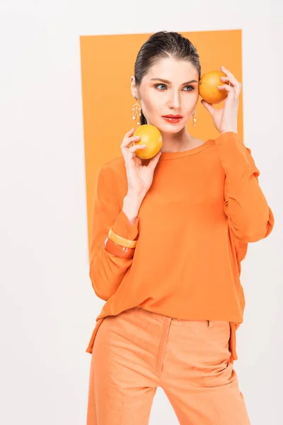 Hermosa mujer elegante sosteniendo naranjas, mirando hacia otro lado y posando con cúrcuma en el fondo - foto de stock