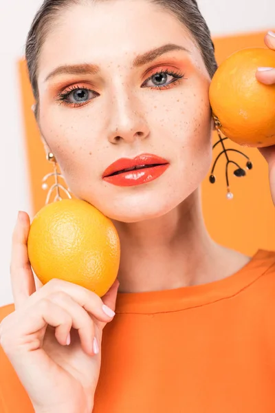 Hermosa mujer elegante sosteniendo naranjas, mirando a la cámara y posando con cúrcuma en el fondo - foto de stock