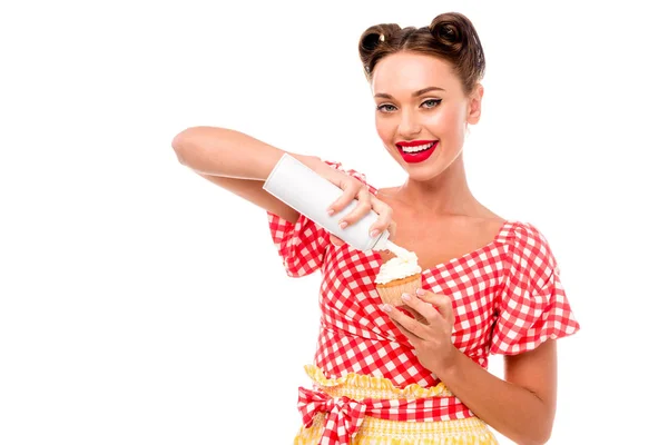 Bella pin up ragazza applicando panna montata su cupcake e guardando la fotocamera isolata su bianco — Foto stock