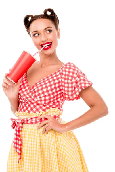 Smiling pin up girl drinking from paper cup with straw and keeping hand on hip isolated on white — Stock Photo