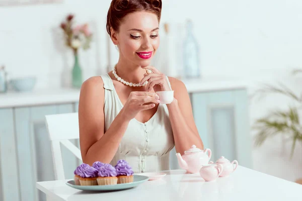 Pritty pin up menina segurando xícara de café enquanto sentado à mesa com prato de cupcakes — Fotografia de Stock