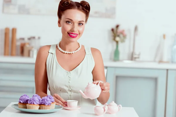 Elegantes lächelndes Pin-up-Mädchen, das am Küchentisch sitzt und in die Kamera blickt — Stockfoto