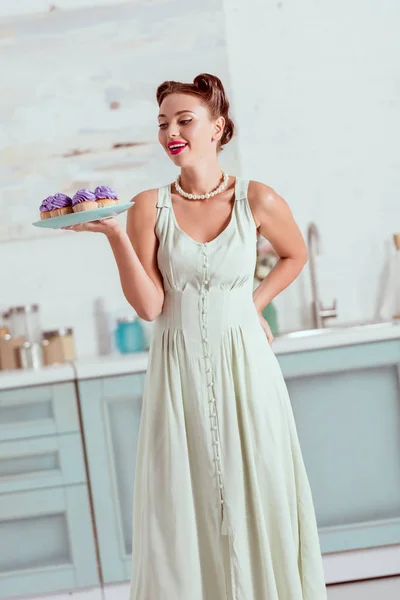 Menina pin up elegante em vestido longo segurando prato cheio de cupcakes caseiros — Fotografia de Stock