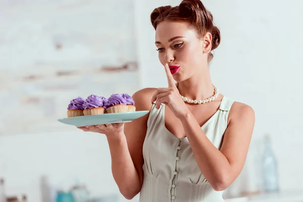 Smiling pin up girl holding plate of cupcakes and showing silence sign — Stock Photo