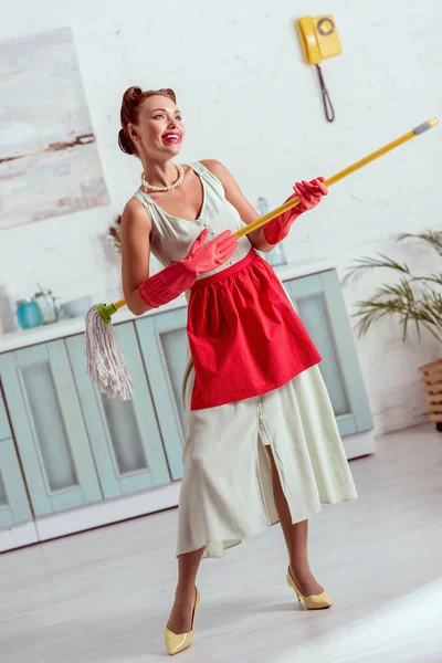 Linda chica pin up en delantal rojo y guantes de goma roja bailando con fregona amarilla - foto de stock