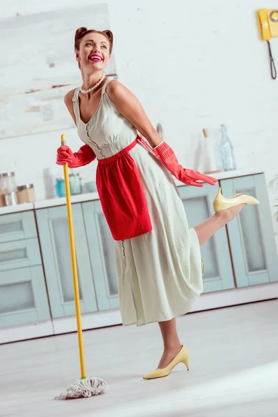 Sorrindo pin up menina em avental vermelho e luvas de borracha vermelha dançando com esfregona amarela — Fotografia de Stock