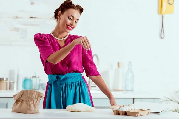 Smiling pin up girl in crimson dress and blue apron adding flour to dough — Stock Photo