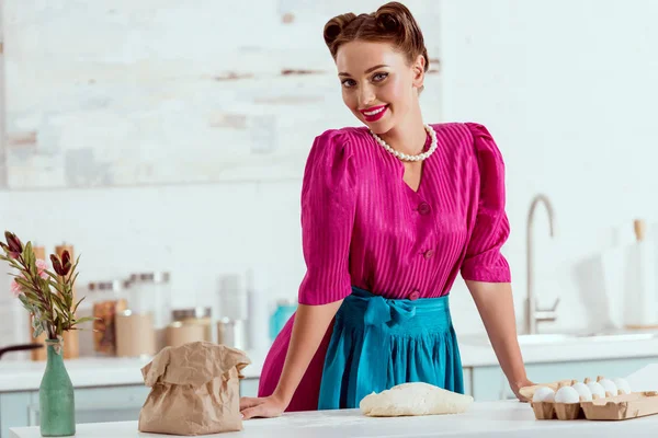 Sorrindo pin up menina em vestido carmesim e avental azul em pé pela mesa da cozinha — Fotografia de Stock