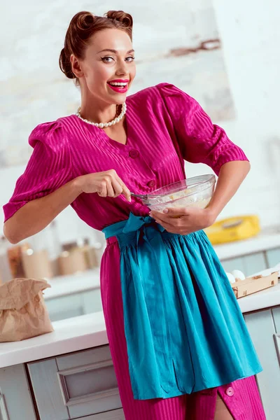 Smiling pin up girl in crimson dress and blue apron mixing ingredients for dough — Stock Photo