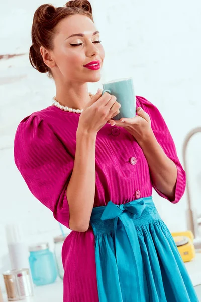 Sorrindo pin up menina em vestido carmesim e avental azul segurando xícara de café com olhos fechados — Fotografia de Stock