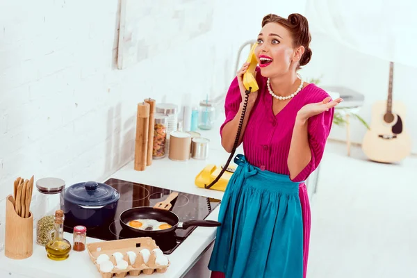 Excited pin up girl talkin on vintage yellow phone while frying eggs — Stock Photo