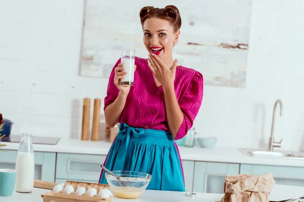 Pretty smiling pin up girl holding glass of milk standing near kitchen table with different products — Stock Photo