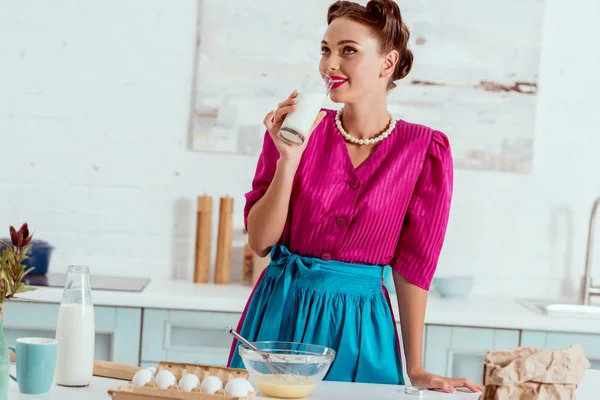 Элегантная булавка девушка пьет молоко стоя возле кухонного стола с различными продуктами — стоковое фото