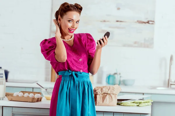 Beautiful pin up girl holding face powder and improving hairstyle while standing near kitchen table with various products — Stock Photo