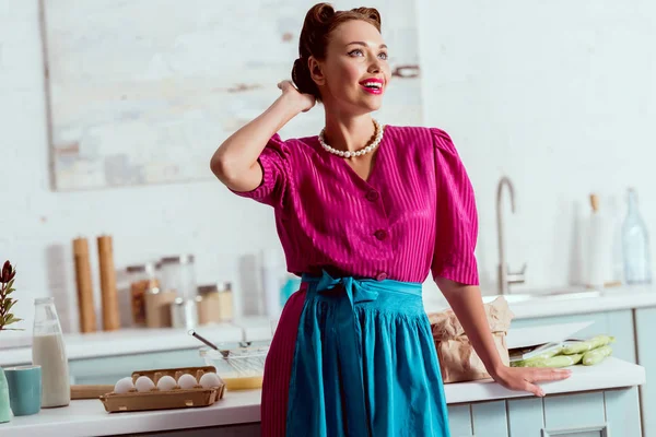 Hermosa chica pin up mejorar el peinado mientras está de pie cerca de la mesa con diferentes productos - foto de stock