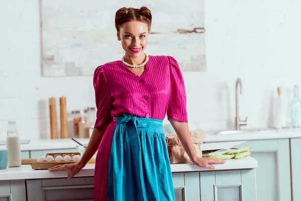 Hermosa chica pin up de pie en la cocina de color azul claro cerca de la mesa con diferentes productos y mirando a la cámara - foto de stock