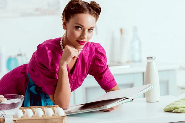 Pretty pin up girl reading recipes book while leaning on kitchen table — Stock Photo