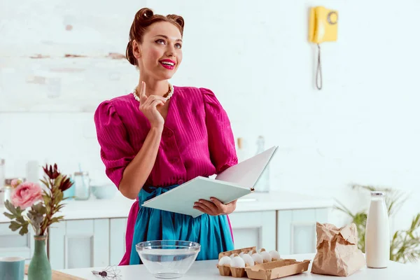 Sorrindo pin up menina segurando livro de receitas e mostrando sinal de ideia — Fotografia de Stock