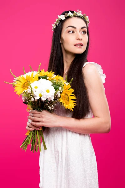 Bella donna bruna in abito bianco e corona floreale tenendo bouquet di fiori e guardando lontano isolato sul rosa — Foto stock