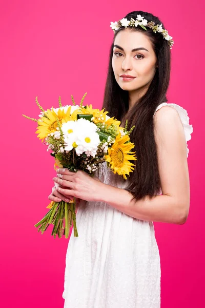 Belle femme brune en robe blanche et couronne florale tenant bouquet de fleurs et regardant la caméra isolée sur rose — Photo de stock