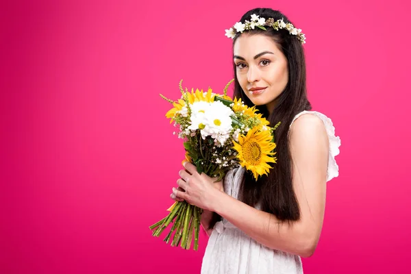 Bella giovane donna bruna in abito bianco e corona floreale tenendo bouquet di fiori e guardando la fotocamera isolata sul rosa — Foto stock
