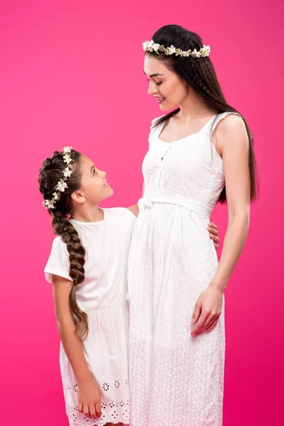 Hermosa madre feliz e hija en vestidos blancos y coronas florales abrazándose y sonriéndose aislados en rosa - foto de stock