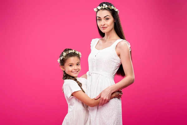 Hermosa madre feliz e hija en vestidos blancos y guirnaldas florales abrazando y sonriendo a la cámara aislada en rosa - foto de stock