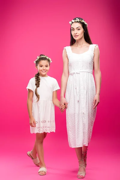Hermosa madre feliz e hija en vestidos blancos y guirnaldas florales tomados de la mano y sonriendo a la cámara en rosa - foto de stock