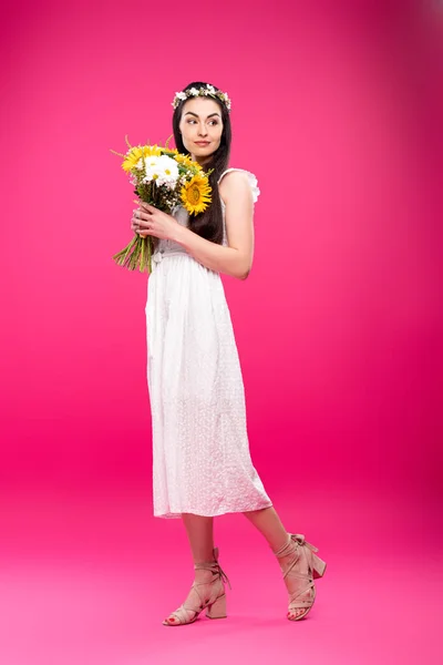 Vue pleine longueur de belle femme brune en robe blanche et couronne florale tenant bouquet sur rose — Photo de stock