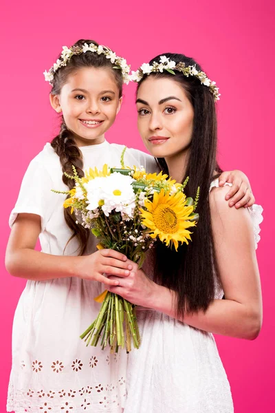 Hermosa madre feliz e hija en vestidos blancos sosteniendo ramo de flores y sonriendo a la cámara aislada en rosa - foto de stock