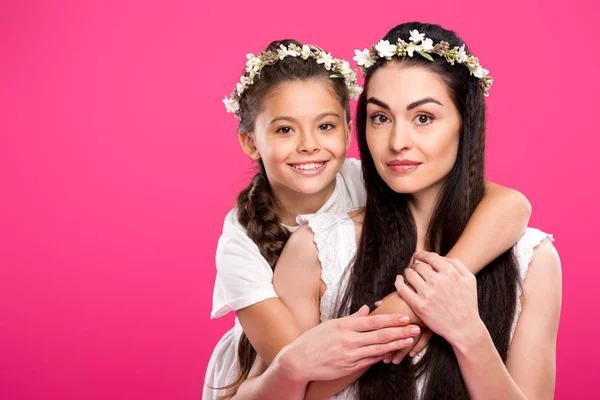 Hermosa madre e hija en vestidos blancos y guirnaldas florales abrazando y sonriendo a la cámara aislada en rosa - foto de stock