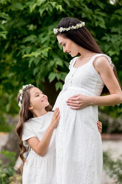 Feliz madre embarazada y linda hija sonriente en vestidos blancos y guirnaldas de pie juntos y sonriendo el uno al otro en el parque - foto de stock