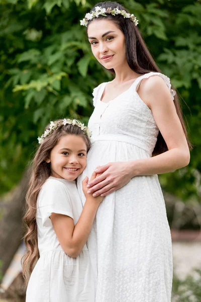 Adorable happy child hugging pregnant mother and smiling at camera in park — Stock Photo