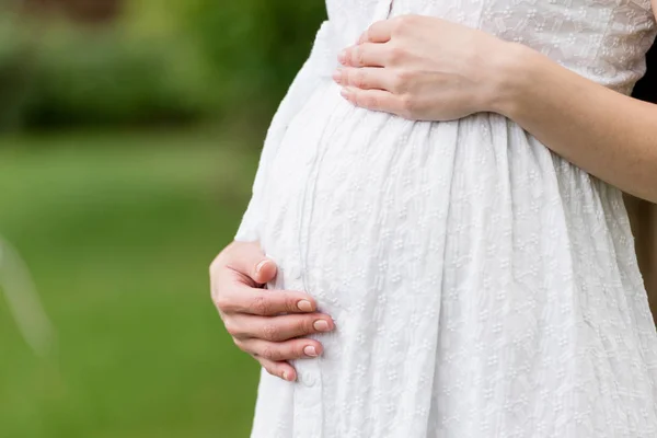 Vista parcial de la mujer embarazada en vestido blanco tocando el vientre mientras está de pie en el parque - foto de stock