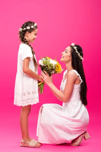 Side view of adorable smiling daughter presenting beautiful flowers to happy young mother on pink — Stock Photo