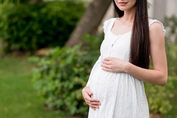 Recortado disparo de sonriente mujer embarazada en vestido blanco de pie en el parque - foto de stock