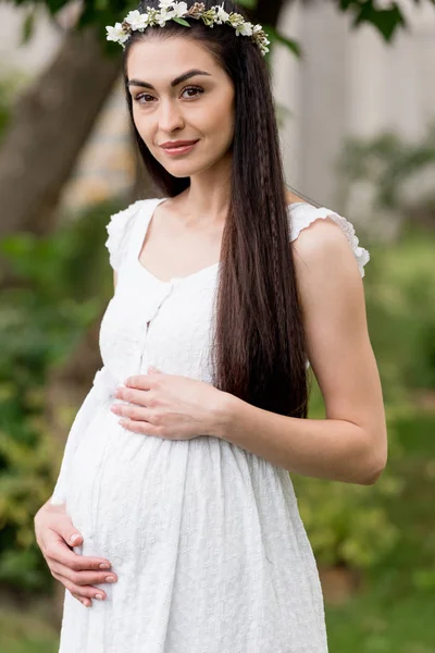 Hermosa mujer embarazada feliz joven en vestido blanco y corona floral sonriendo a la cámara en el parque - foto de stock