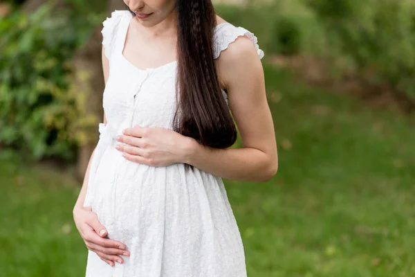 Tiro cortado de mulher grávida em vestido branco tocando barriga enquanto em pé no parque — Fotografia de Stock