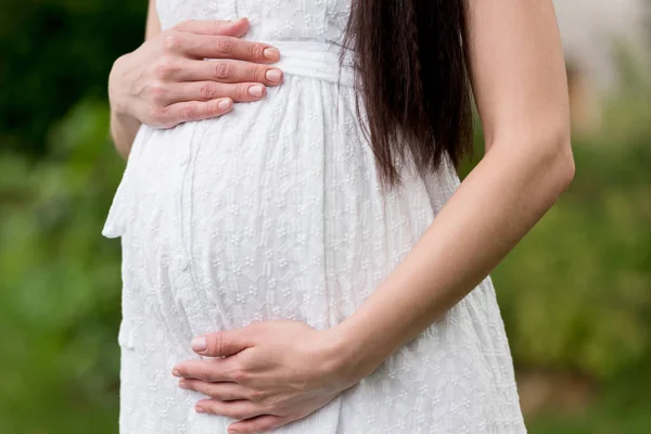 Mid section of pregnant woman in white dress touching belly while standing in park — Stock Photo